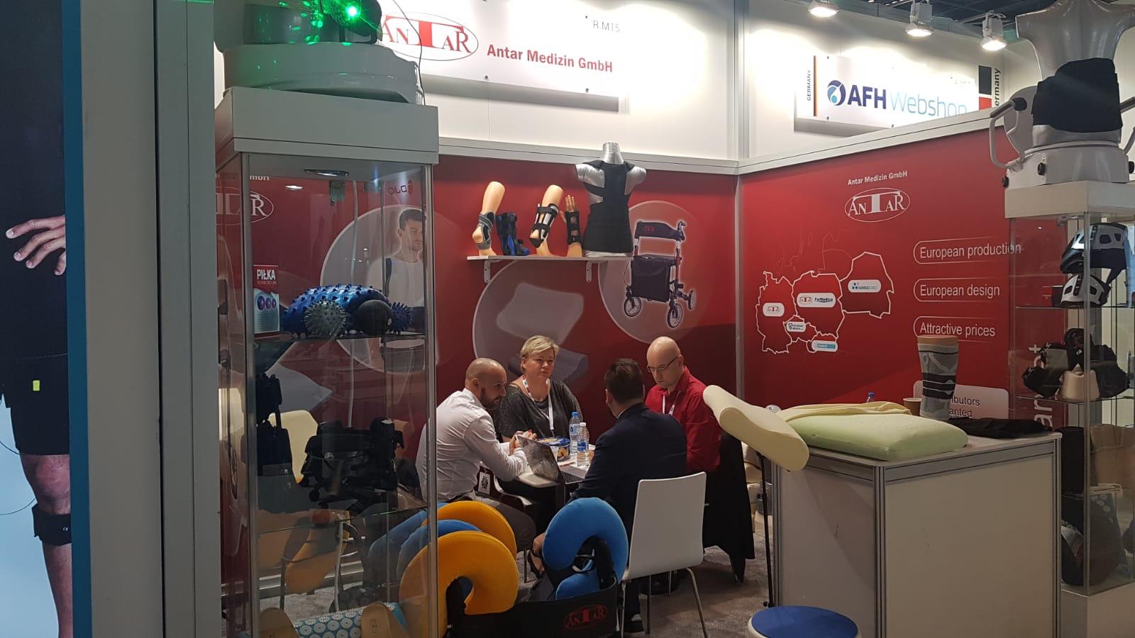 ANTAR Messestand auf der Dubai 2020 Messe – Präsentation von orthopädischen Hilfsmitteln und Rehaprodukten. Moderne Ausstellung mit Prothesen, Bandagen, Rollatoren und ergonomischen Kissen. Geschäftsbesprechung mit Fachleuten vor einer roten Markenwand mit europäischen Produktions- und Designinformationen.