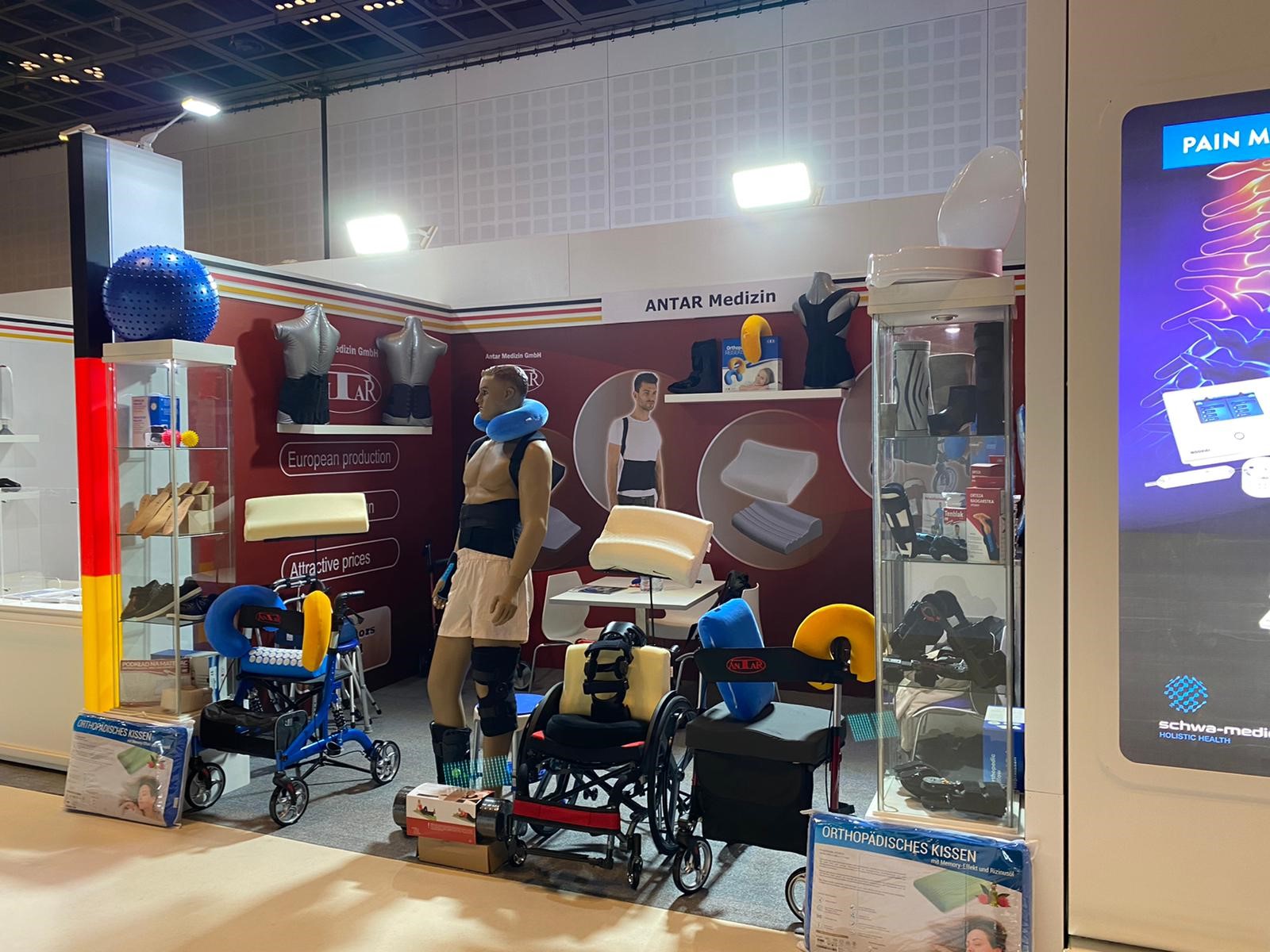 Messestand der Firma **ANTAR Medizin** auf der **Arab Health 2021** in Dubai. Der Stand präsentiert eine Vielzahl orthopädischer Hilfsmittel, darunter Rollstühle, Rollatoren, orthopädische Kissen und Bandagen. In der Mitte steht eine **lebensgroße Schaufensterpuppe**, die eine Rückenbandage und eine Knieorthese trägt. Auf einem **blauen Rollator** vorne sind ergonomische **Nackenkissen** in Blau und Gelb befestigt. Ein **roter Rollstuhl** mit Rückenstützkissen und Beinlagerungshilfe steht daneben. Links eine **Glasvitrine** mit orthopädischen Einlagen, Kompressionsstrümpfen und Fitnessbällen. An der Rückwand sind Produktbilder von **orthopädischen Kissen** und Rückenstützen zu sehen. Eine zweite Glasvitrine auf der rechten Seite zeigt Bandagen, Orthesen und medizinische Hilfsmittel. Im Hintergrund befinden sich ein kleiner **Besprechungstisch** mit Stühlen und ein Plakat mit dem Schriftzug „**European production - European design - Attractive prices**“. Der Messestand ist in den Farben **Rot, Weiß und Schwarz** gestaltet und beleuchtet.